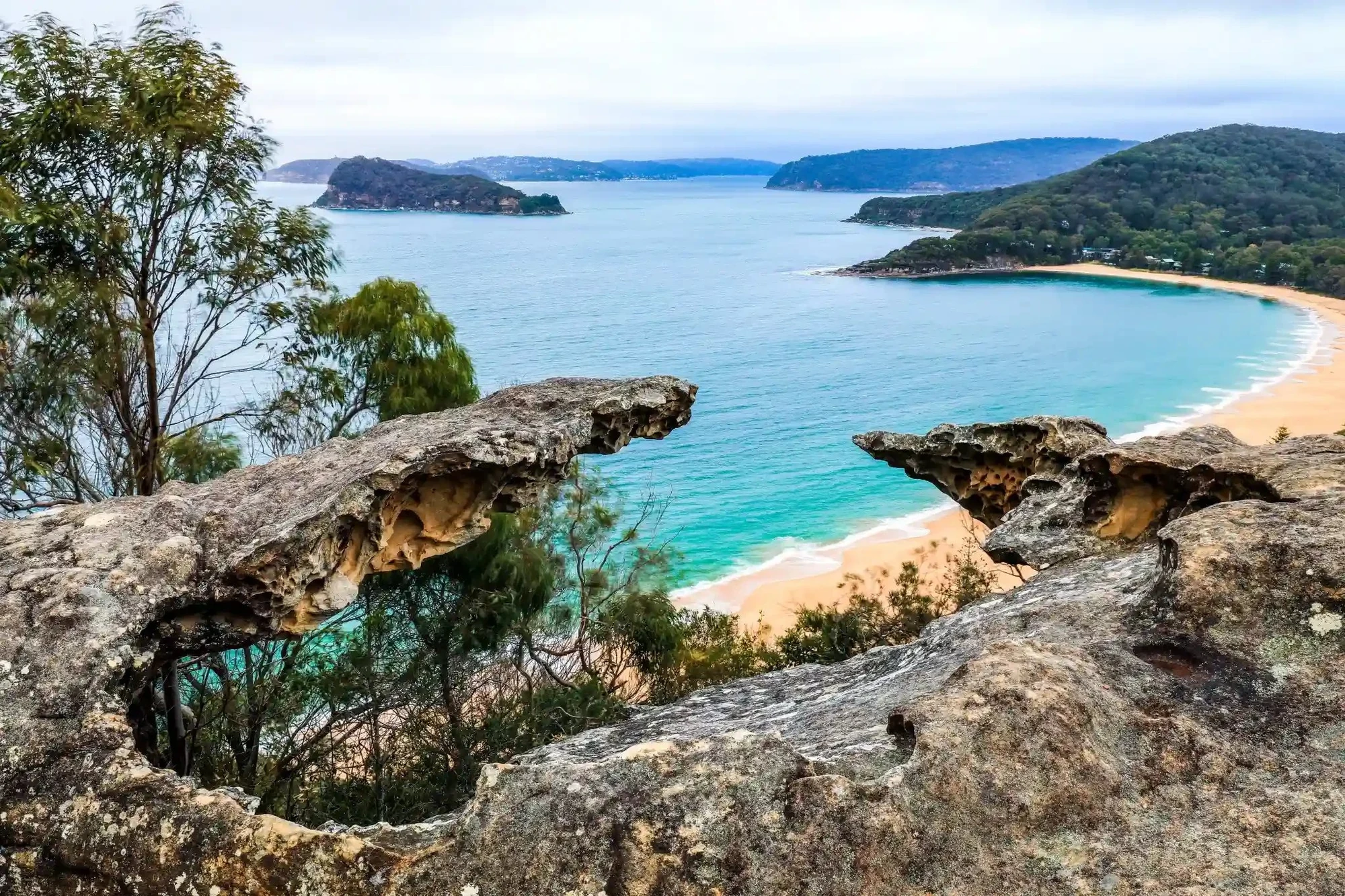 View from a rocky lookout over turquoise water at Pearl Beach on NSW's Central Coast, with tree-covered headlands in the distance